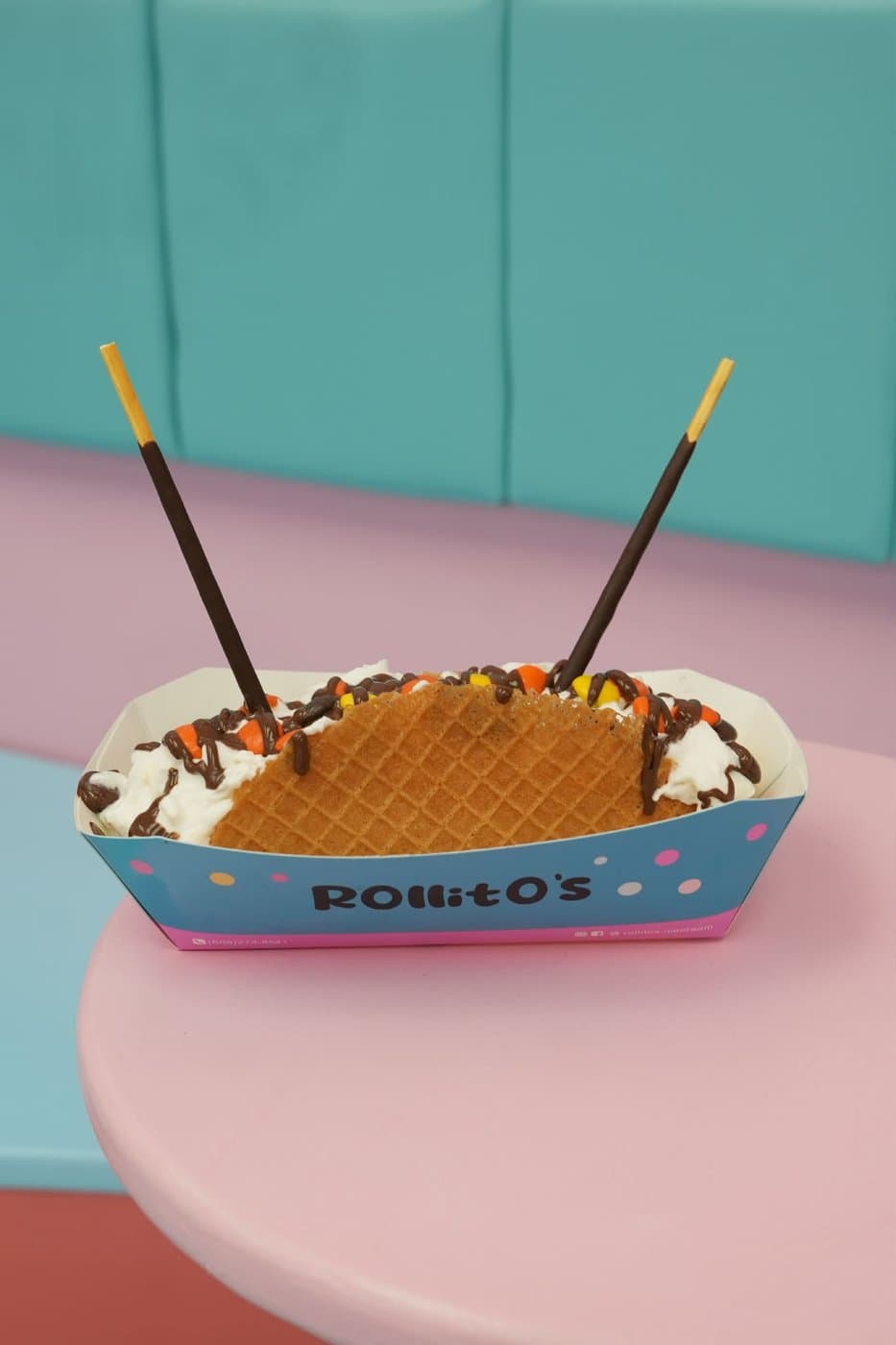 Golden waffle taco roll with whipped cream, peanut butter candies, chocolate drizzle, and Pocky sticks, on a pink table with a teal backdrop