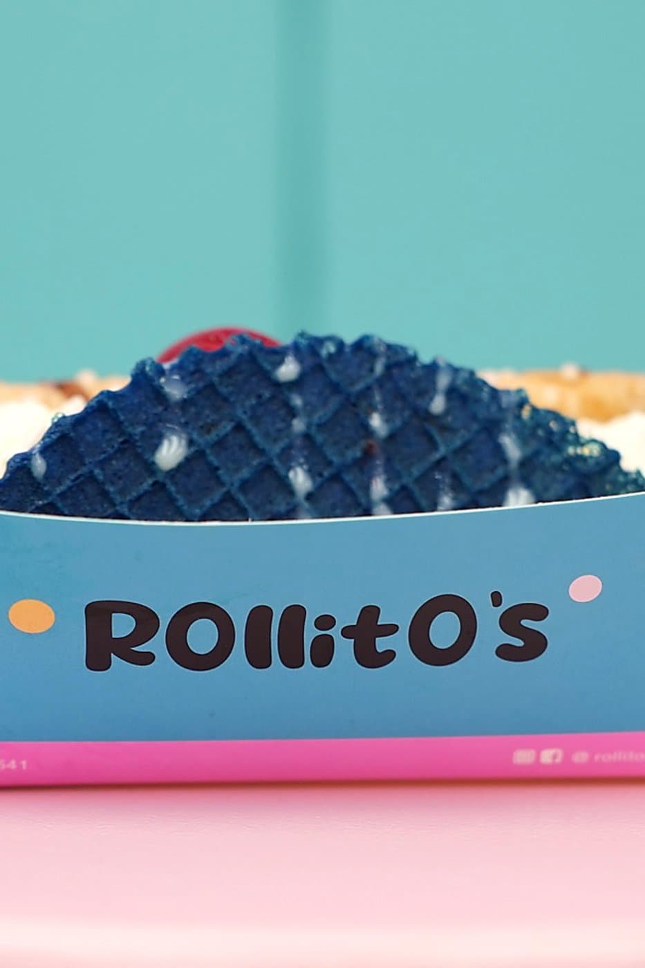 Blue waffle taco roll with whipped cream, wafer rolls, and cherries on top, on a pink table with a teal backdrop